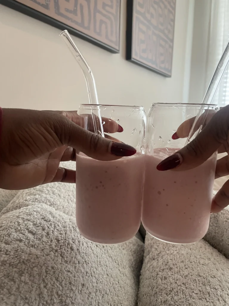 Two hands clinking pale-pink smoothies in cozy living room setting.