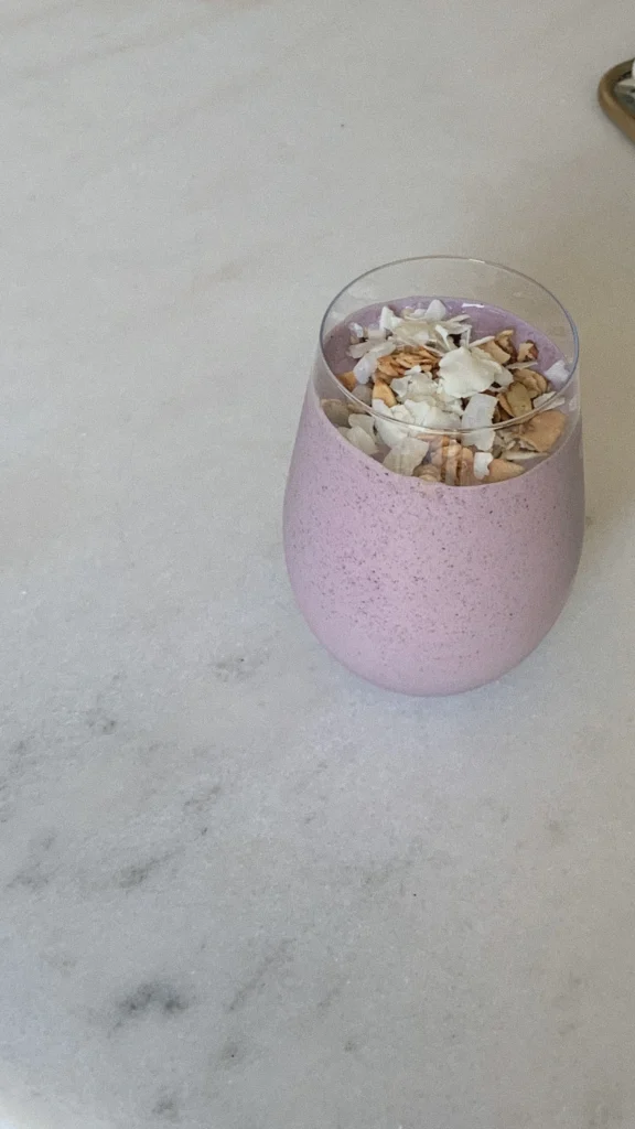 Lilac smoothie in curved glass topped with coconut and granola.