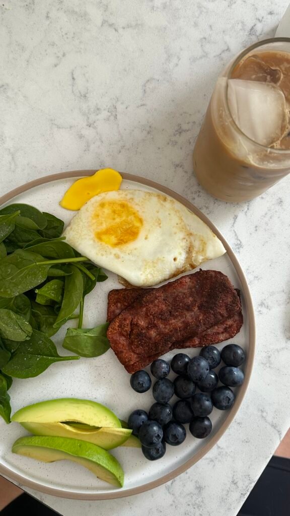Breakfast plate with fried egg, seared lean meat, fresh spinach, avocado, and blueberries.